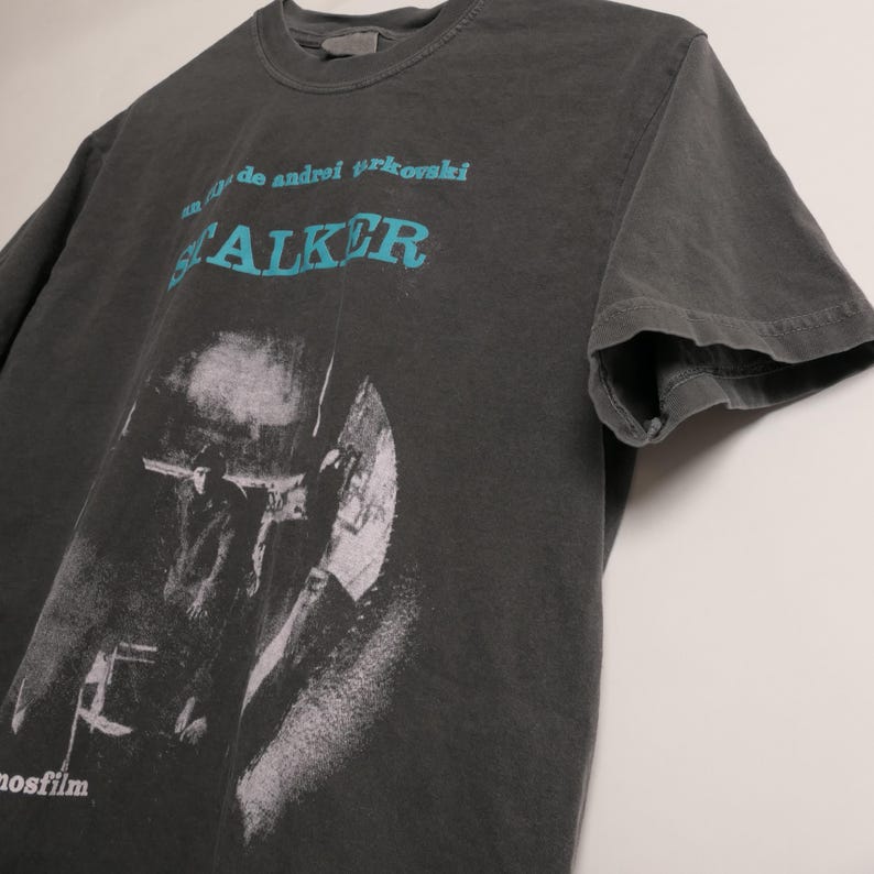 May include: A dark gray t-shirt with a black and white image of a person holding a gun. The text "film by Andrei Tarkovsky STALKER" is printed in blue on the shirt.