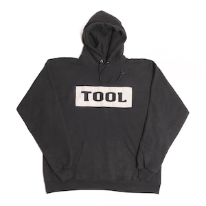 May include: A black hooded sweatshirt with a white rectangular design on the front. The word "TOOL" is printed in bold, black letters within the white rectangle. The sweatshirt has a drawstring hood and a front pocket.