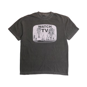 Peut inclure: T-shirt gris foncé avec un motif d'écran de télévision affichant les mots "WATCH TV" au-dessus de deux personnes. L'image est en noir et blanc. Le t-shirt est à col rond.