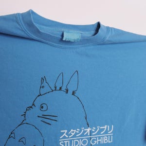 May include: A light blue t-shirt featuring a black and white illustration of a cartoon character with large ears and the text "STUDIO GHIBLI" in Japanese. The shirt has a crew neck and short sleeves.