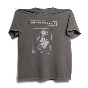 Peut inclure: Un t-shirt gris foncé avec une image en noir et blanc d'un visage et le texte "THIS MORTAL COIL" imprimé sur le devant.