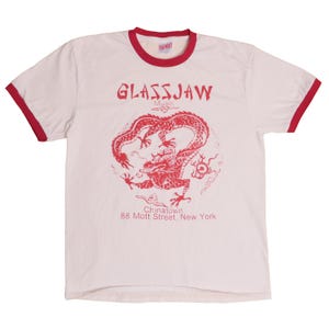 以下が含まれることがあります： 赤色のトリムが襟と袖に施されたクリーム色のTシャツ。前面には、赤いドラゴンのイラストの上に「GLASSJAW MUSIC」の文字が描かれています。ドラゴンの下には「Chinatown 88 Mott Street, New York」と書かれています。