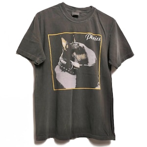 Może przedstawiać: Ciemnoszary t-shirt z krótkim rękawem z nadrukiem graficznym przedstawiającym bulteriera w obroży z ćwiekami w żółtej kwadratowej ramce. Słowo Pixies jest nadrukowane białą kursywą w prawym górnym rogu grafiki.