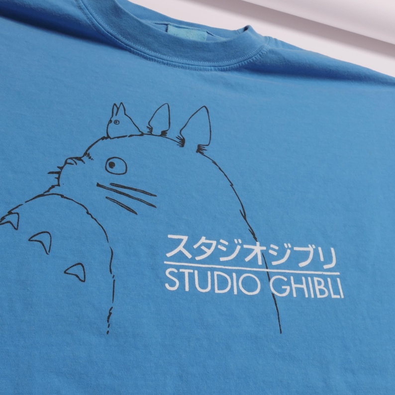 May include: A blue t-shirt featuring a black and white illustration of a cartoon character with the text "スタジオジブリ STUDIO GHIBLI" in white. The shirt has a crew neck and short sleeves.