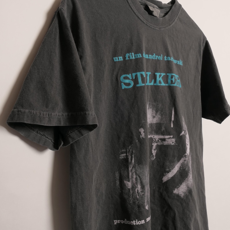 May include: A dark gray t-shirt with a faded print of a movie poster. The poster features the text "un film andrei tarkovski" and "STALKER" in blue. The poster also includes a black and white image of a scene from the movie.