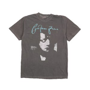 May include: A charcoal grey short-sleeve t-shirt featuring a light blue script logo that reads Cocteau Twins above a high-contrast, grainy black and white photographic portrait of a person's face centred on the front of the garment.