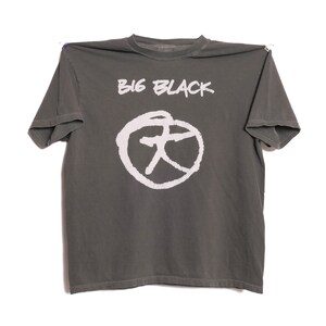以下が含まれることがあります： 白の「BIG BLACK」の文字と白い円形グラフィックがプリントされた、グレーの半袖Tシャツ。柔らかい素材で作られています。白い背景にTシャツが表示されています。