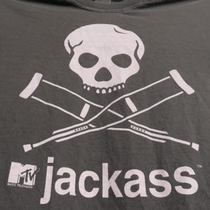 K&ouml;nnte beinhalten: Dunkelgraues T-Shirt mit wei&szlig;em Totenkopf und gekreuzten Kr&uuml;cken. Darunter steht "jackass" mit dem MTV-Logo. Das Shirt besteht aus einem weichen Material.