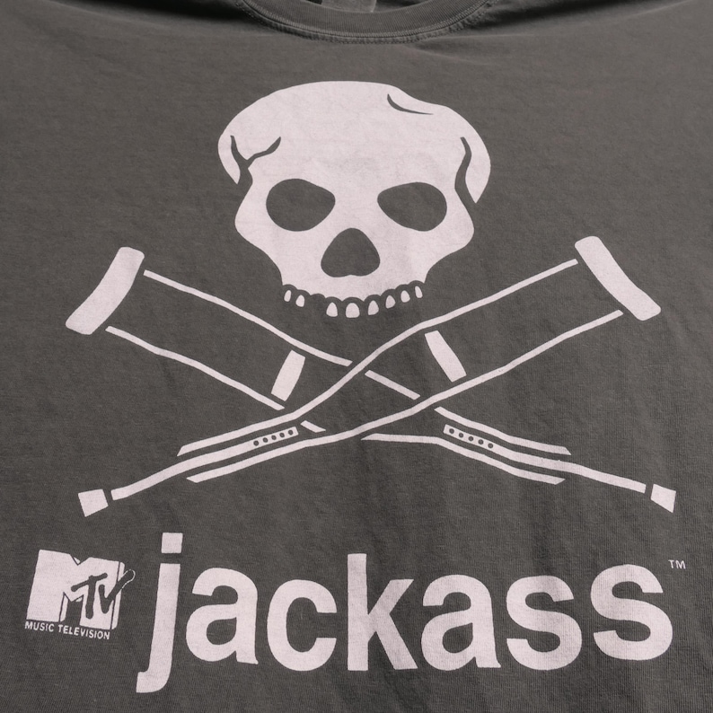 K&ouml;nnte beinhalten: Dunkelgraues T-Shirt mit wei&szlig;em Totenkopf und gekreuzten Kr&uuml;cken. Darunter steht "jackass" mit dem MTV-Logo. Das Shirt besteht aus einem weichen Material.
