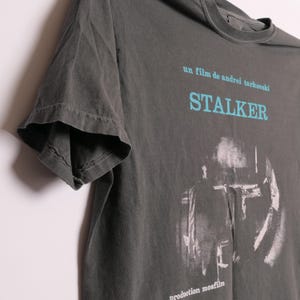 May include: A dark gray t-shirt with a faded image of a scene from the film "Stalker" by Andrei Tarkovsky. The text "un film de andrei tarkovsky" and "STALKER" are printed in blue on the shirt. The text "production mosfilm" is printed in white at the bottom of the shirt.