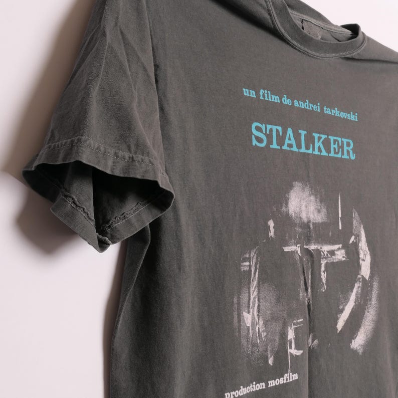 May include: A dark gray t-shirt with a faded image of a scene from the film "Stalker" by Andrei Tarkovsky. The text "un film de andrei tarkovsky" and "STALKER" are printed in blue on the shirt. The text "production mosfilm" is printed in white at the bottom of the shirt.