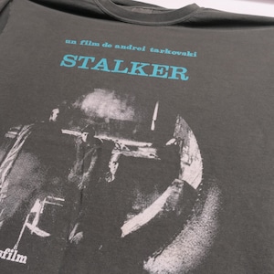 May include: A dark gray t-shirt with a black and white image of three people in a room. The text "un film de andrei tarkovski STALKER" is printed in blue on the shirt.