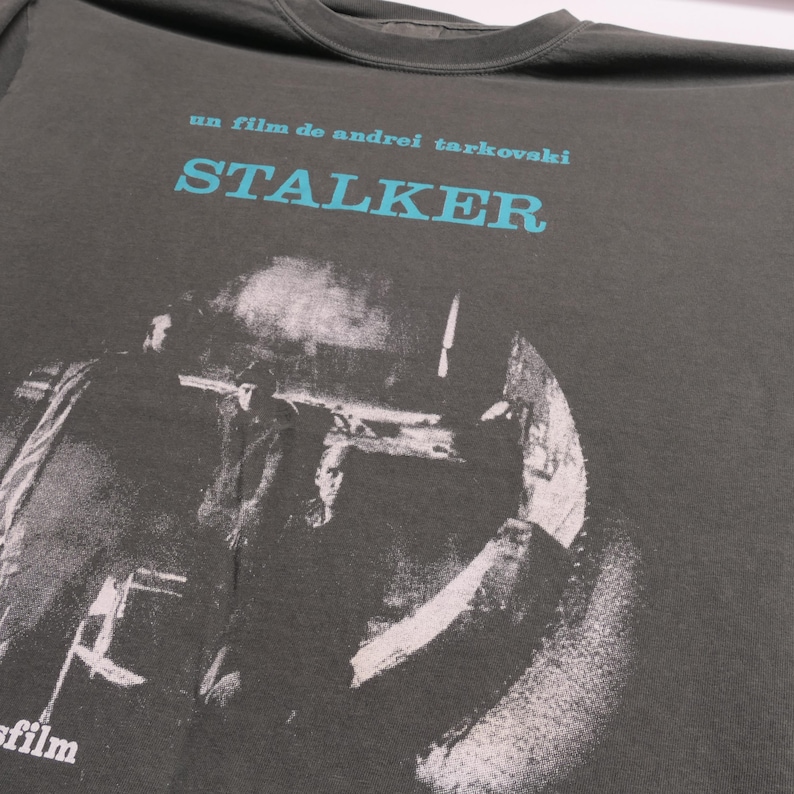 May include: A dark gray t-shirt with a black and white image of three people in a room. The text "un film de andrei tarkovski STALKER" is printed in blue on the shirt.
