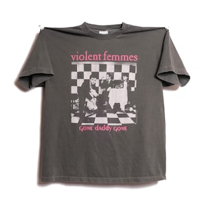 May include: Dark gray t-shirt featuring the text "violent femmes" in pink above a black and white image of a band. The text "GONE daddy GONE" is printed below the image. The shirt is made of a soft material.