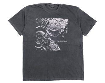 May include: Dark gray t-shirt featuring a white graphic of ammonite fossils. The text "The Sundays" is printed below the graphic. The shirt has short sleeves and a crew neck.