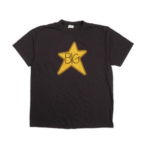 Puede incluir: Una camiseta negra de manga corta con un gráfico grande de una estrella amarilla de cinco puntas centrado en el pecho. Dentro de la estrella, la palabra BIG está impresa en una fuente negra y audaz de estilo dibujado a mano. Cuello redondo.