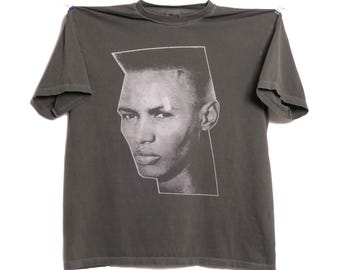 Pode incluir: Camiseta cinza com um retrato em preto e branco de uma pessoa com um corte de cabelo reto. O retrato é delineado em branco. A camiseta está pendurada em um varal contra um fundo branco.