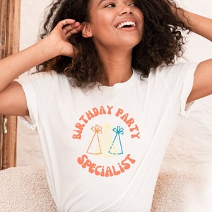 Op de afbeelding: Een wit T-shirt met een kleurrijke afbeelding waarop "Verjaardagsfeest-specialist" staat met twee feestmutsen. De afbeelding bevindt zich in een cirkel met een perzikkleurige achtergrond.
