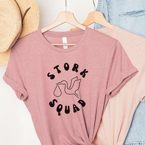 Op de afbeelding: Een roze T-shirt met een zwarte afbeelding van een ooievaar die een baby in een luier draagt. De afbeelding is omringd door de tekst "Stork Squad".