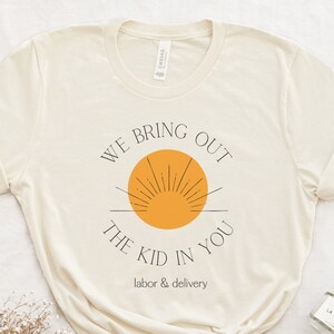 May include: A beige t-shirt with the text "We bring out the kid in you. Labor & delivery" and a stylized orange sun graphic.