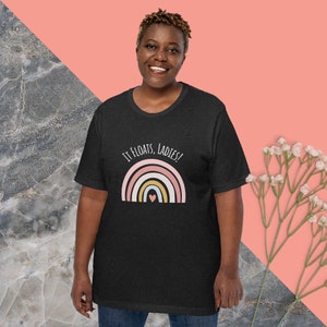 May include: Dark gray t-shirt with the text "IT FLOATS, LADIES!" above a rainbow design with a heart. The rainbow features pink, yellow, and blue arches. The shirt is short-sleeved.