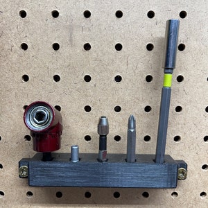 May include: A black plastic tool holder with five slots for holding various tools, including a red and silver right-angle drill attachment, a silver drill bit, a silver screwdriver bit, a silver screwdriver bit, and a silver magnetic screwdriver bit. The holder is mounted on a pegboard.