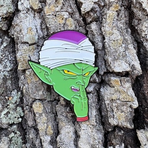 May include: A green enamel pin featuring the character Piccolo from Dragon Ball Z. The pin shows Piccolo with a white turban, purple head, and yellow eyes. His finger is raised to his nose. The pin is set against a textured tree bark background.