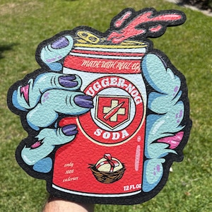 May include: A novelty sign shaped like a soda can, featuring the text "UGGER-NOG SODA" and a cross symbol. The can is held by a pair of zombie hands with blue skin and purple nails. The sign also includes the text "MADE WITH REAL EGG" and "only 1000 calories".