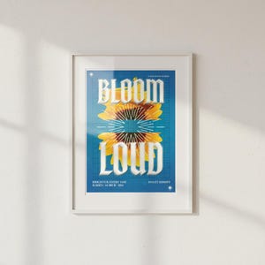 May include: Framed art print with a sunflower design and the words "BLOOM LOUD" in white on a blue grid. The print also includes the text "BRIGHTER EVERY DAY" and "PINO25 SERIES03". The artwork is in a white frame.