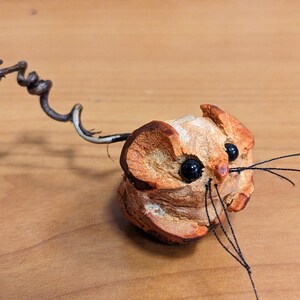 May include: A small, brown, wooden mouse figurine with black eyes and a wire tail. The mouse has a long, thin, black wire whiskers.