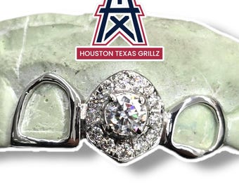 May include: A silver-colored grill with a large, central, clear gemstone surrounded by smaller stones. The grill is set on a light green mold. The text "HOUSTON TEXAS GRILLZ" is displayed above the grill.