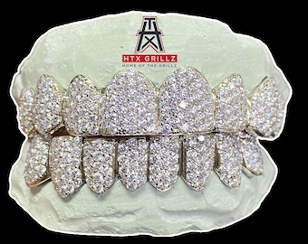 May include: A set of gold grillz with a full diamond design. The grillz are set in a mold of a mouth. The text "HTX GRILLZ HOME OF THE GRILLZ" is visible above the grillz.
