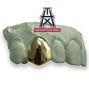 May include: Gold grillz with a single front tooth, set in a green mold. The grillz are shaped to fit the front teeth. The mold is labeled "HOUSTON TEXAS GRILLZ" with a red and blue logo of a derrick.