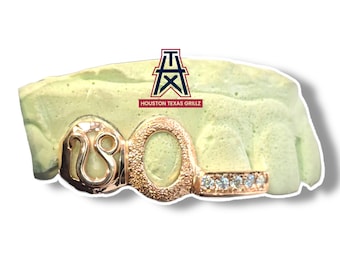 Op de afbeelding: Roségouden grill met de letter "O" en een gestileerd ontwerp. De onderste rij heeft een rij kleine, heldere stenen. Het logo "Houston Texas Grillz" is zichtbaar op de achtergrond.