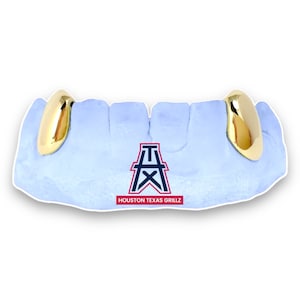 May include: Gold-colored tooth grillz, displayed on a light blue dental mold. The grillz are highly polished and reflect light. The mold has a logo with the text "HOUSTON TEXAS GRILLZ".