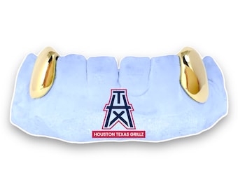 May include: Gold-colored tooth grillz, displayed on a light blue dental mold. The grillz are highly polished and reflect light. The mold has a logo with the text "HOUSTON TEXAS GRILLZ".