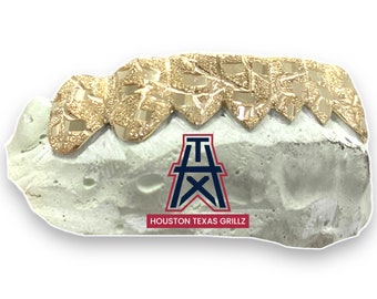 May include: Gold-colored grillz with a textured surface, resting on a white mold. The logo for Houston Texas Grillz is visible below the grillz. The grillz are designed to fit over the top teeth.