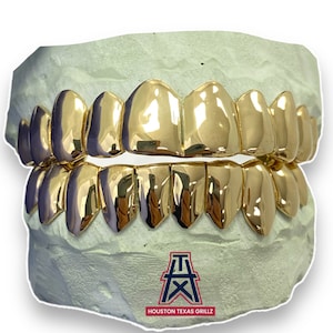 May include: Gold grillz for teeth, with a design of pointed teeth. The grillz are displayed in a mold, with the logo "HOUSTON TEXAS GRILLZ" at the bottom.