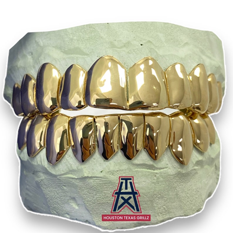 May include: Gold grillz for teeth, with a design of pointed teeth. The grillz are displayed in a mold, with the logo "HOUSTON TEXAS GRILLZ" at the bottom.