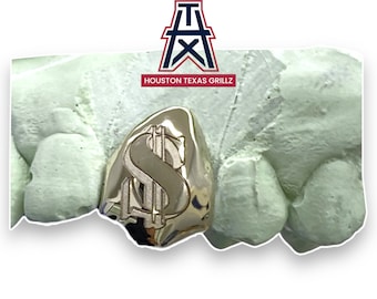 May include: A gold-colored tooth grill with a dollar sign design. The grill is on a white mold. The text "HOUSTON TEXAS GRILLZ" is visible above the grill.