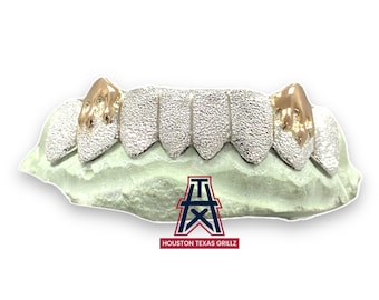May include: Silver-colored grillz with gold-colored fangs, displayed on a light green mold. The grillz have a textured surface. The Houston Texas Grillz logo is visible at the bottom.