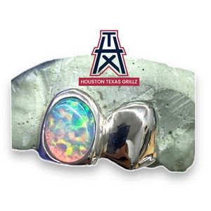 May include: A close-up of a silver tooth grill with two opal stones. The opal stones are iridescent with shades of pink, green, and blue. The grill is set on a gray mold. The text "HOUSTON TEXAS GRILLZ" is visible below the grill.