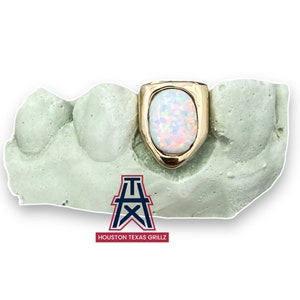 May include: A gold tooth grill with a single opal stone in the front. The grill is set in a mold for display. The text "HOUSTON TEXAS GRILLZ" is visible below the mold.