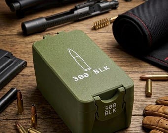 May include: An olive green ammunition box with the text "300 BLK" and a bullet graphic. The box is surrounded by disassembled firearm parts, bullets, and a gun cleaning kit. The items are arranged on a wooden surface.