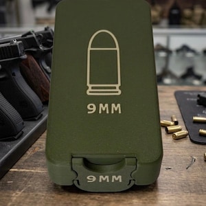 May include: An olive green ammunition case with a white outline of a bullet and the text "9MM" printed on the front. The case is closed and has a hinged opening. The text "9MM" is also printed on the bottom of the case.