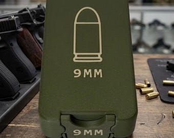 May include: An olive green ammunition case with a white outline of a bullet and the text "9MM" printed on the front. The case is closed and has a hinged opening. The text "9MM" is also printed on the bottom of the case.