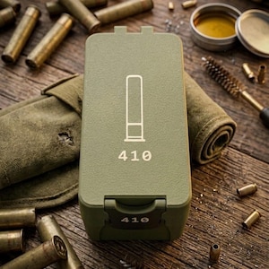 May include: An olive green ammunition box with the number "410" printed on it. The box has a white graphic of a shell. The image also shows spent shell casings, a cleaning brush, and a small container of lubricant, all on a wooden surface.