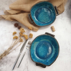 Puede incluir: Dos cuencos de cerámica hechos a mano con un vibrante esmalte turquesa y asas de color marrón oscuro. Los cuencos están colocados sobre una superficie blanca con un paño de arpillera, cubiertos y flores secas. También se ve un corcho.