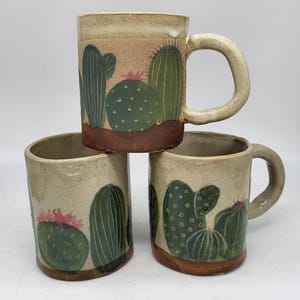 Puede incluir: Tres tazas de cerámica con acabado beige moteado y base marrón. Cada taza presenta cactus verdes pintados a mano con flores rosas. Las tazas tienen forma cilíndrica, un asa resistente y una superficie texturizada con pinceladas visibles.