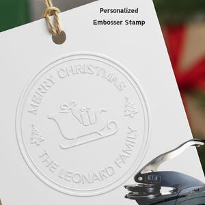 以下が含まれることがあります： 白いエンボス加工のクリスマスカード。テキスト「MERRY CHRISTMAS」と「THE LEONARD FAMILY」がそりを囲んでいます。カードは紐付きのタグに付いています。黒とシルバーのエンボッサーが右下にあります。